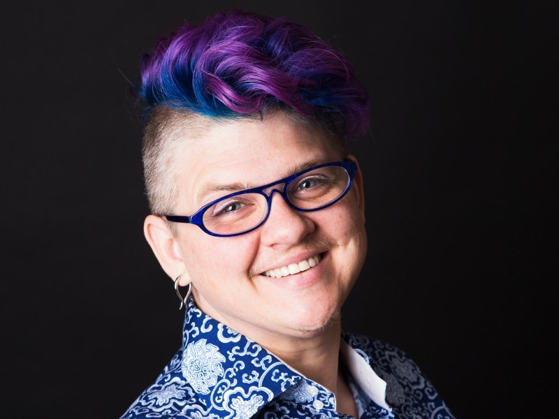 Headshot of Frances Reed, a white, non-binary person with a bright purple and blue mohawk. They are wearing glasses and a patterned blue and white shirt and smiling into the camera.