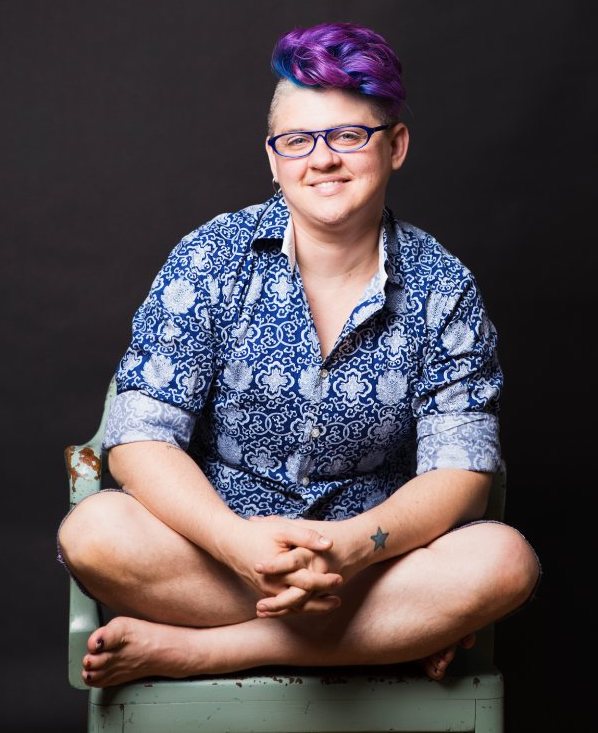 Photo of Frances Reed, a white, non-binary person with a bright purple and blue mohawk. They are wearing glasses and a patterned blue and white shirt and smiling into the camera. They are sitting cross-legged on a chair with their hands folded.