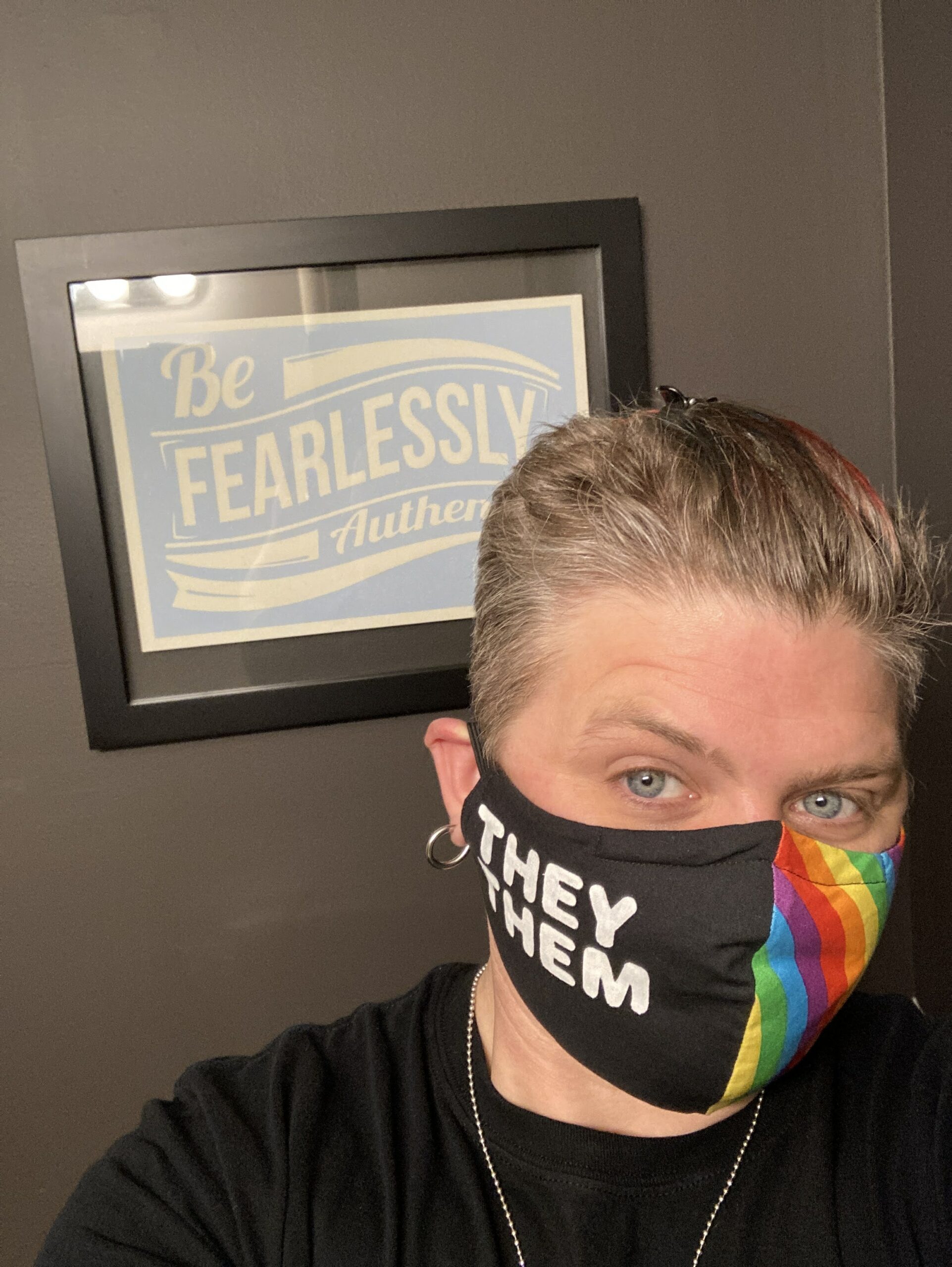 Photograph of Frances wearing a rainbow face mask that reads "they  them" in front of a framed photo that says "fearlessly authentic"